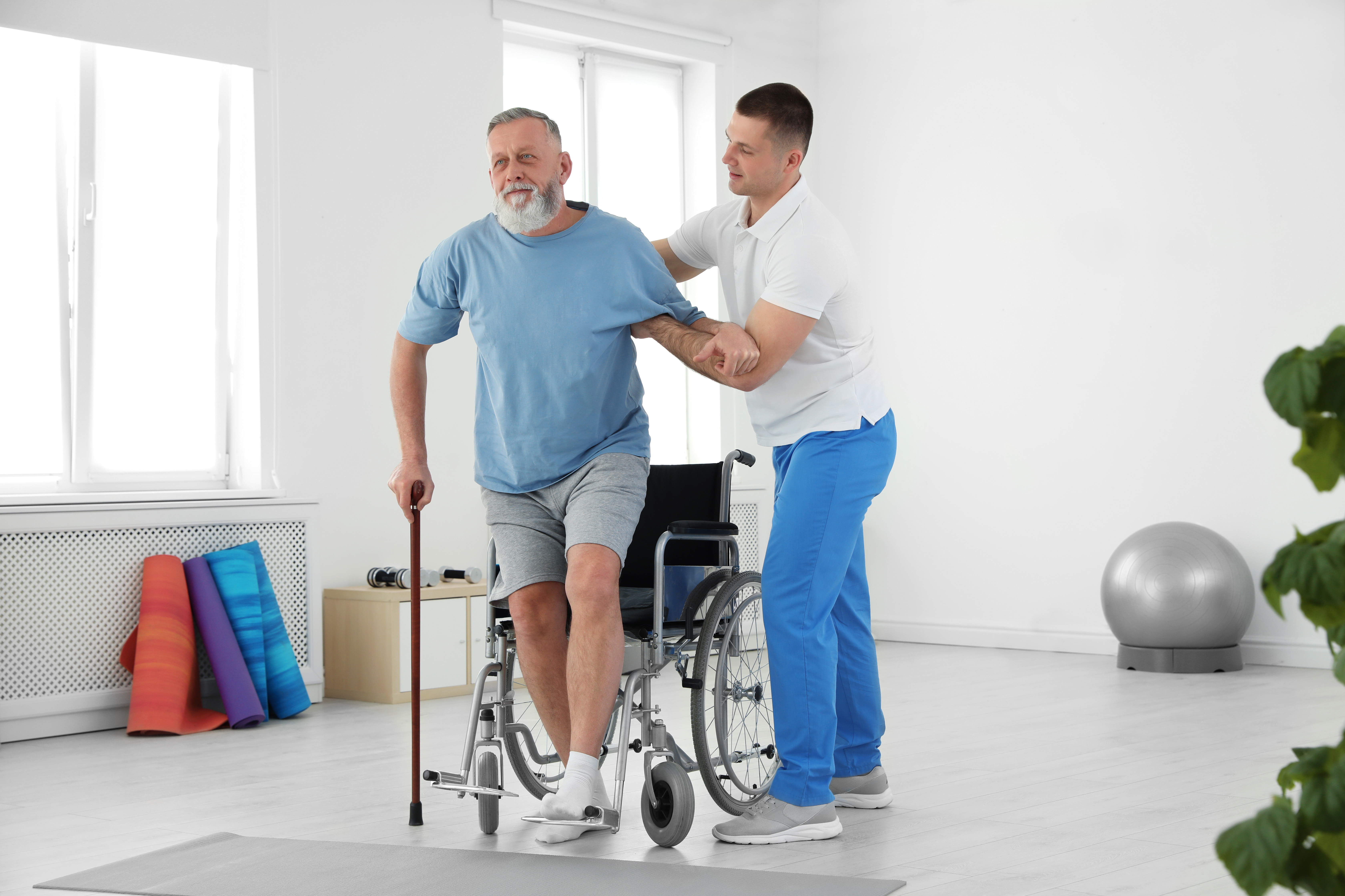 an older man stands from a wheelchair with the assistance of a younger physical therapist 