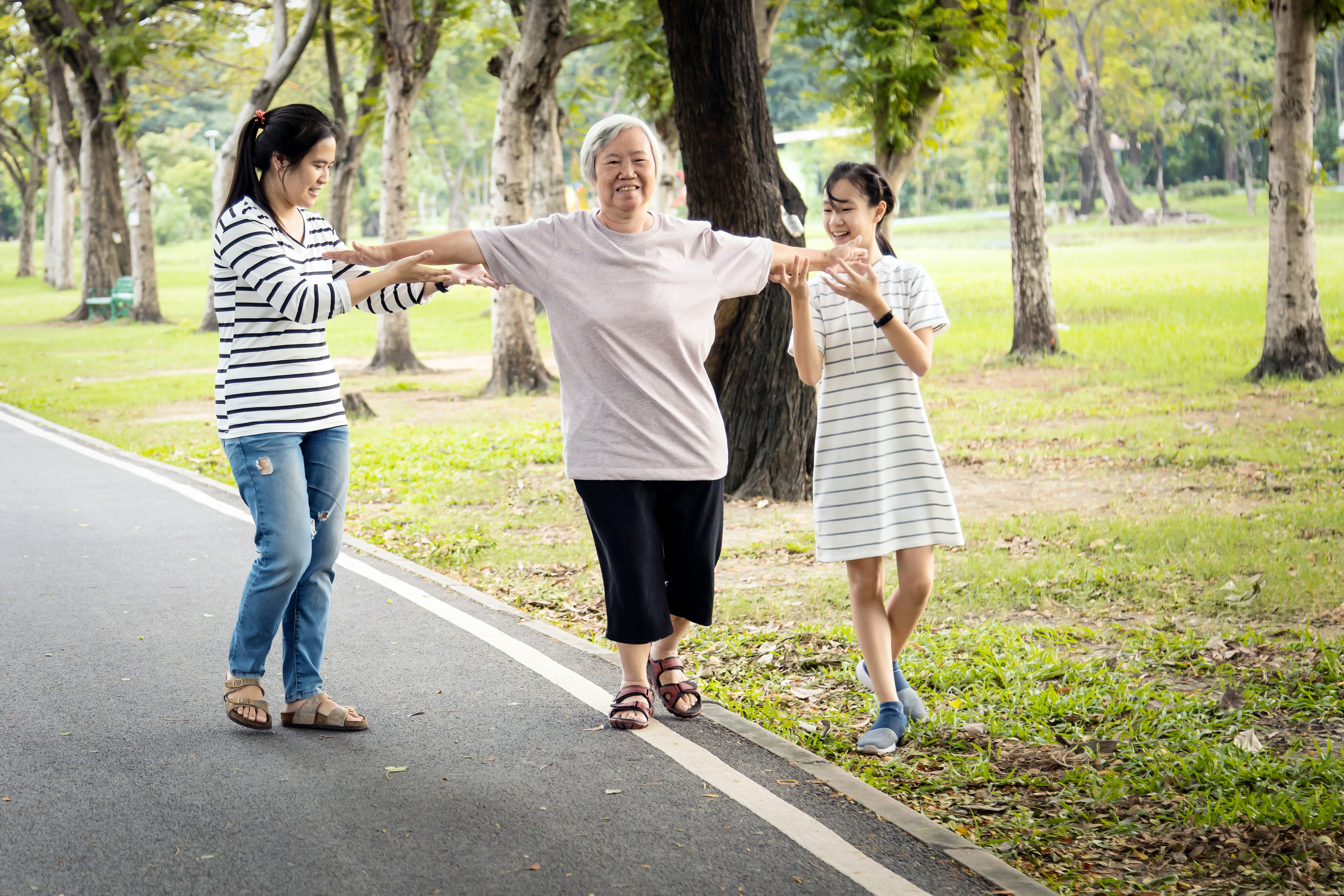 Three asian women walking together outside, a grandmother, mother, and daughter. The grandmother is balancing on a white line, testing for fall risk