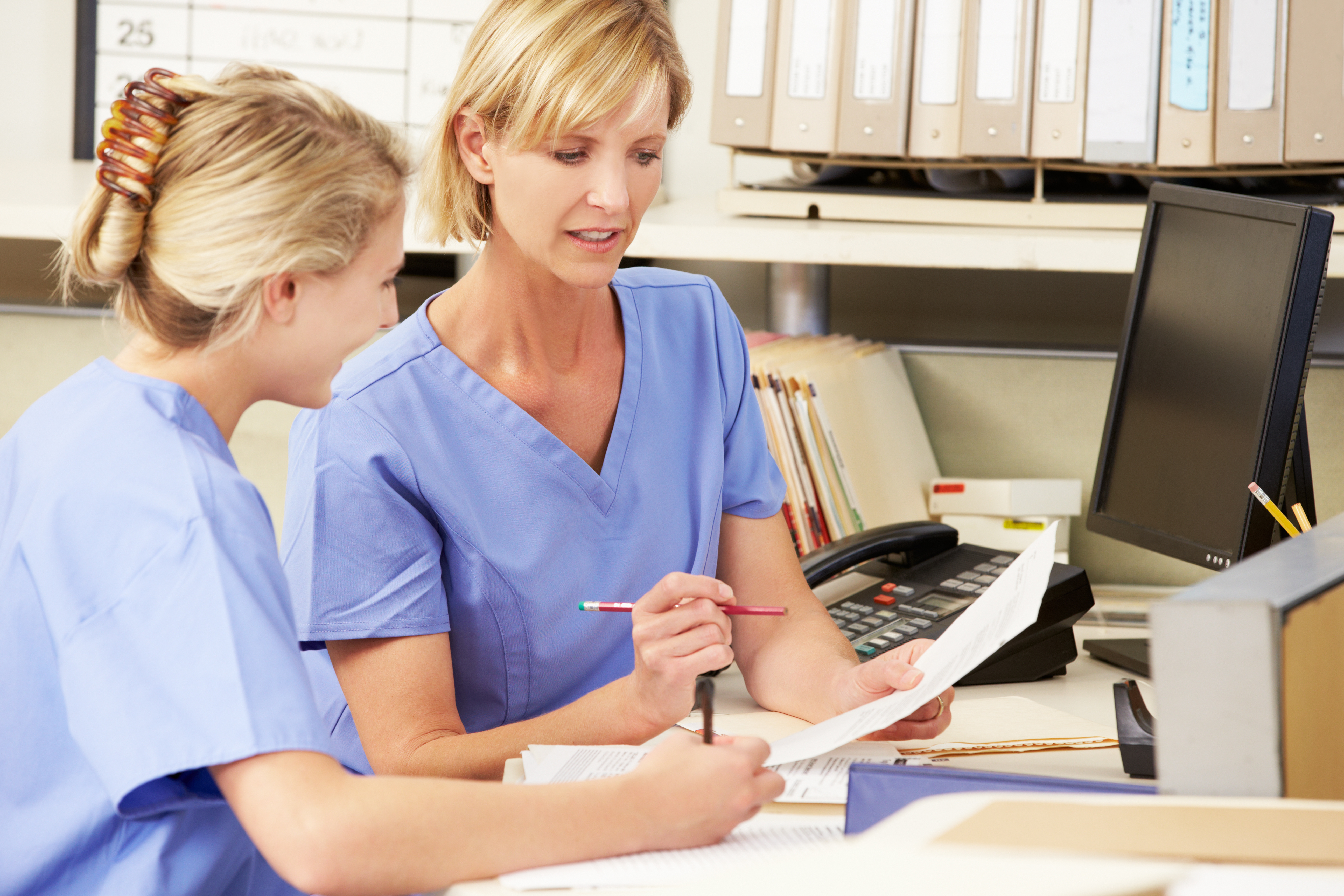 two nurses reviewing paperwork together at a computer desk 