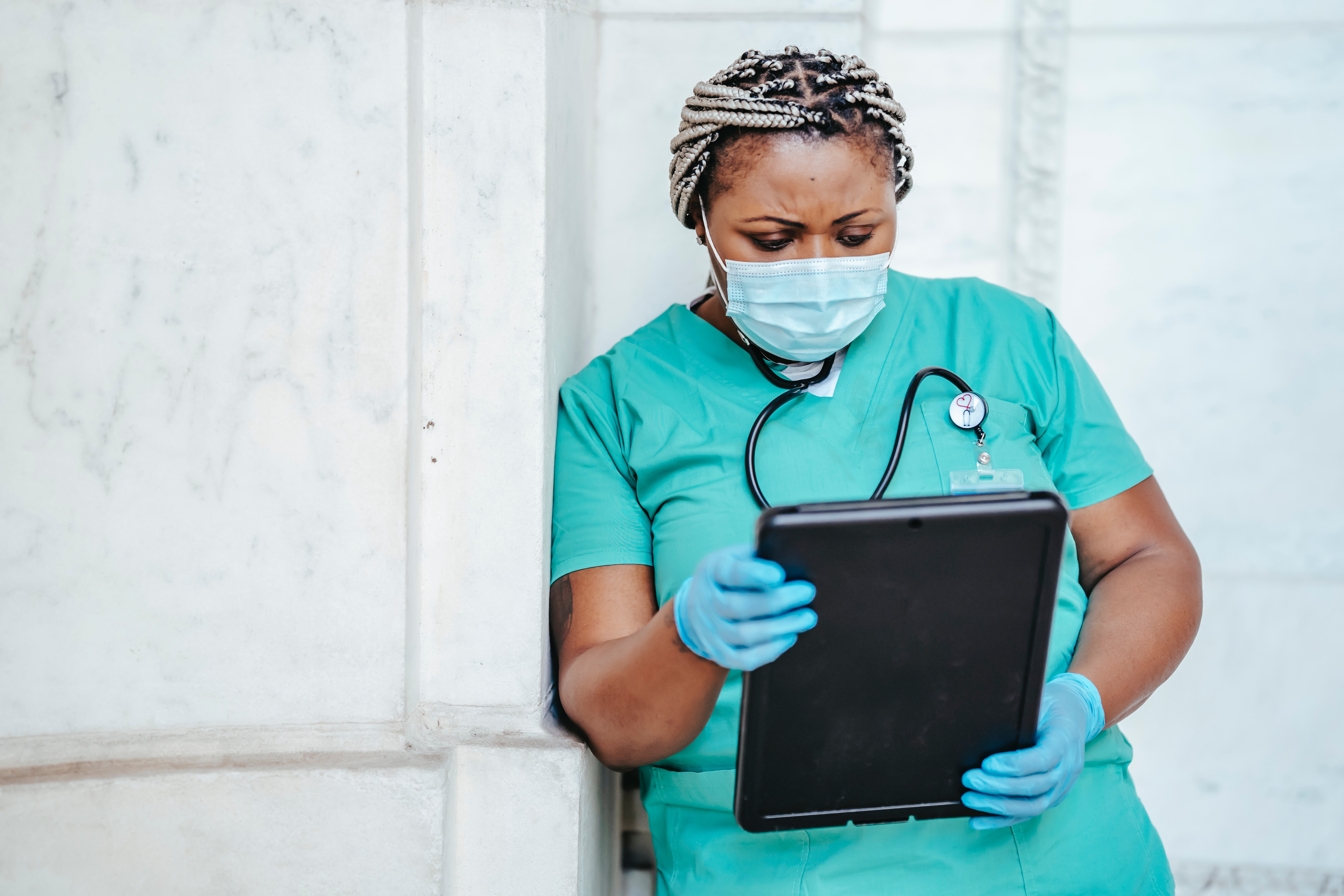 a nurse leans against a wall, brow furrowed, as she reads information from a tablet