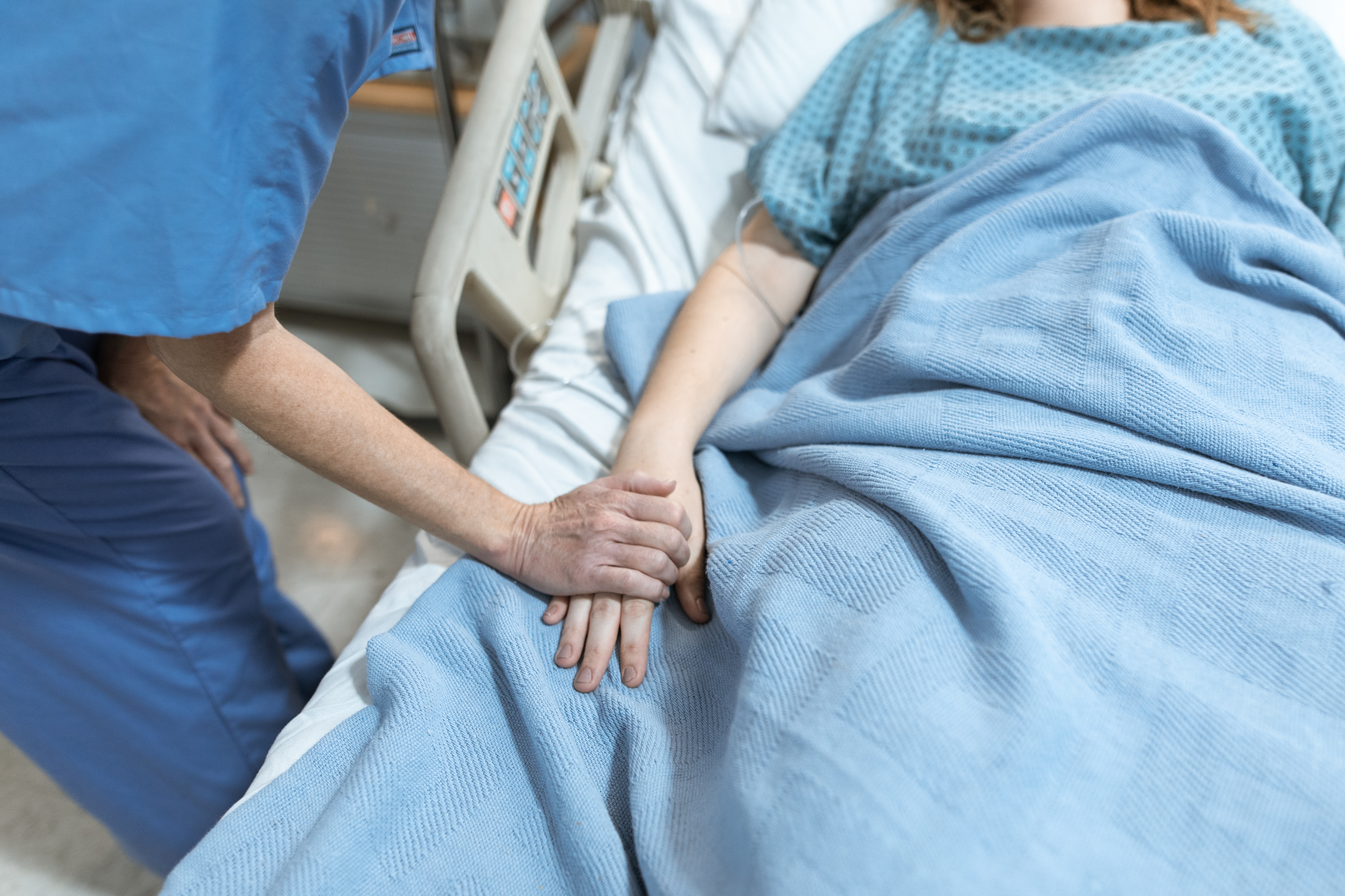 a nurse in blue scrubs holds the hand of a patient in a hospital bed. 